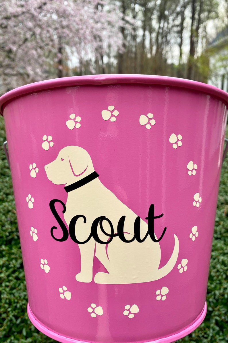 May include: Pink metal dog treat bucket with a white dog silhouette and the name "Scout" in black lettering. The bucket is decorated with white paw prints.