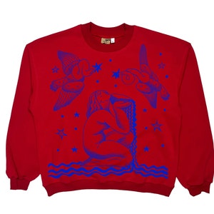 Könnte beinhalten: Roter Pullover mit einem blauen Grafikdruck einer Frau mit langen Haaren, zwei Vögeln und Sternen. Die Grafik erinnert an ein Vintage-Tattoo-Design.