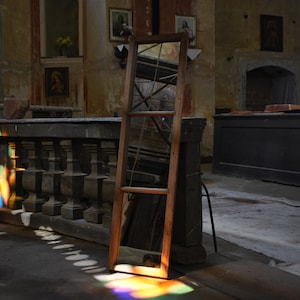 May include: A tall, rectangular mirror with a natural wood frame, divided into three vertical sections by horizontal wooden bars. The mirror reflects the surrounding interior space and is illuminated by a patch of vibrant, multicoloured light.