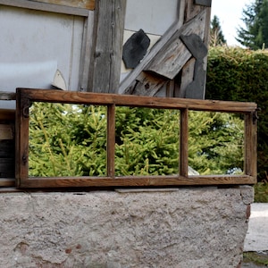 Könnte beinhalten: Ein rechteckiger, dreiteiliger Holzfensterrahmen mit dunkelbraunem Finish. Der Rahmen steht vor einer verwitterten, strukturierten Betonmauer, mit einem grünen, belaubten Hintergrund, der durch die Scheiben sichtbar ist. Ein rustikales, Vintage-Objekt.