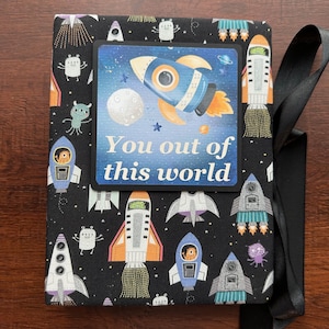 May include: A black fabric-covered journal with a space-themed pattern of rockets, aliens, and planets. A rectangular label on the front reads "You out of this world" with a rocket ship illustration. A black ribbon is tied around the journal.