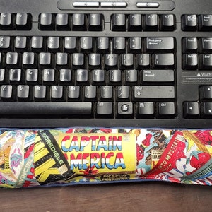 Puede incluir: Un reposamuñecas colorido con un collage de portadas de cómics con personajes como Capitán América, Lobezno y el Increíble Hulk. El reposamuñecas está hecho de tela y tiene un borde azul.