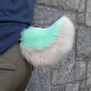Puede incluir: Una cola de piel sintética esponjosa en colores verde menta y crema. La cola está sujeta a la cintura de una persona, sobre un fondo de pared de piedra. Es un accesorio de disfraz.