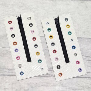 May include: Two white leather zippered pouches with colourful eyelets. Each pouch has a black zipper and a row of eyelets in different colours.