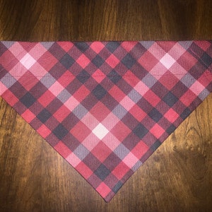 May include: A red and black plaid dog bandana with a white background. The bandana is triangular in shape and has a stitched edge.