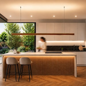 Puede incluir: Cocina moderna con una gran isla, dos taburetes y una lámpara de madera. La cocina cuenta con armarios blancos, una encimera negra y una gran ventana con vistas a un jardín verde. El suelo es de madera.
