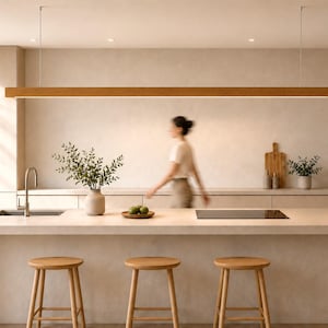 May include: A modern kitchen with light wood cabinetry and a large island with three wooden stools. A long, rectangular wooden light fixture hangs above the island. A person is walking in the background.