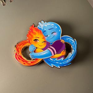 Puede incluir: Un colorido pin de esmalte con dos personajes de dibujos animados abrazándose. Uno es una llama con tonos naranja y rojo, y el otro es una figura azul parecida al agua. El pin tiene forma de símbolo de infinito.