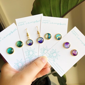 May include: Handmade earrings with gold-coloured settings and abstract designs in teal, blue, purple, and gold. The earrings are displayed on cards with the text "handmade by zoe campbell". The earrings include stud and drop styles.