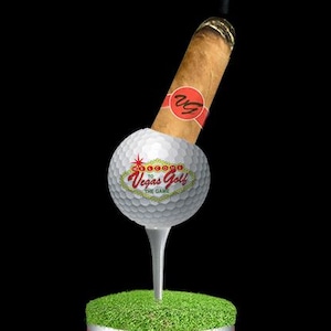 Könnte beinhalten: Ein neuartiger Golf-Tee-Halter mit einem Golfball mit "Vegas Golf"-Branding, der eine brennende Zigarre hält. Der Halter ist eine Dose mit dem Text "Stogie Holder Authentic Golf Ball!" und "Removable Golf Tee!" vor schwarzem Hintergrund.