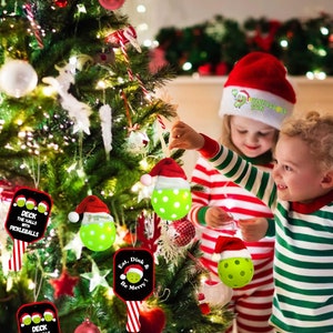 May include: Two children decorate a Christmas tree with pickleball ornaments. The ornaments are green and white with a Santa hat on top. One ornament has the text "Eat, Dink & Be Merry!" and another has the text "Deck the Halls with Pickleballs".