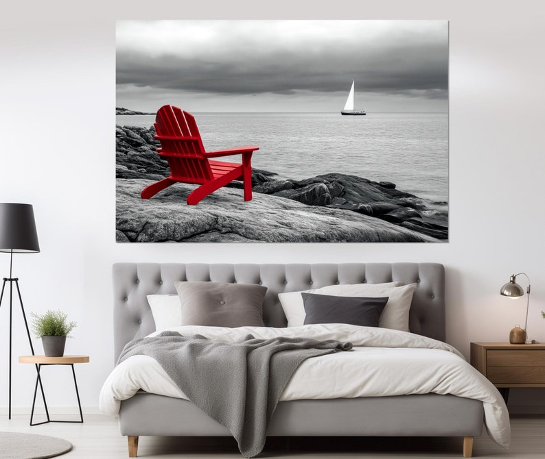 May include: A red Adirondack chair sits on a rocky shore overlooking a body of water with a sailboat in the distance. The sky and water are black and white, while the chair is a bright red.