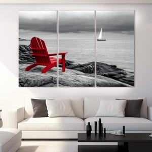 May include: A red Adirondack chair sits on a rocky beach with a sailboat in the distance. The background is black and white, while the chair is a bright red.