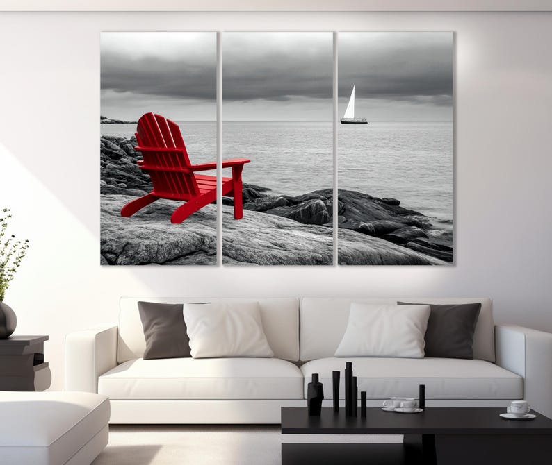 May include: A red Adirondack chair sits on a rocky beach with a sailboat in the distance. The background is black and white, while the chair is a bright red.
