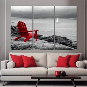 May include: A red Adirondack chair sits on a rocky beach with a sailboat in the distance. The scene is in black and white except for the chair.