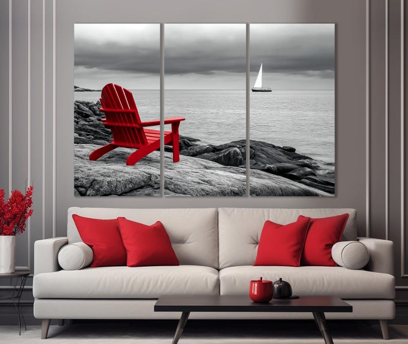 May include: A red Adirondack chair sits on a rocky beach with a sailboat in the distance. The scene is in black and white except for the chair.