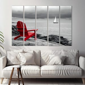 May include: A red Adirondack chair sits on a rocky shore with a sailboat in the distance. The background is a black and white photo, while the chair is in color.