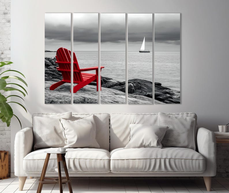 May include: A red Adirondack chair sits on a rocky shore with a sailboat in the distance. The background is a black and white photo, while the chair is in color.