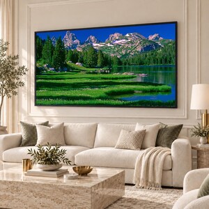 Può includere: Una grande stampa murale rettangolare raffigurante un paesaggio di montagne innevate, una fitta foresta di pini verdi e un lago calmo che riflette il cielo blu, il tutto racchiuso in una sottile cornice nera montata su una parete bianca.