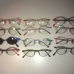 May include: A collection of 12 pairs of eyeglasses in various colors and styles, including round, oval, and rectangular frames. Some frames are made of metal, while others are made of plastic. The eyeglasses are arranged in rows on a white surface. The brand name "SM & CO" is visible on several of the frames.