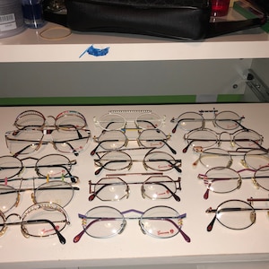 May include: A collection of 14 pairs of eyeglasses with various frame styles and colors, including gold, silver, and black. The eyeglasses are arranged in rows on a white surface.