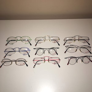 May include: Eight pairs of eyeglasses with different colored frames, including green, blue, silver, and red. The frames are all made of metal and have a round shape. The lenses are clear.