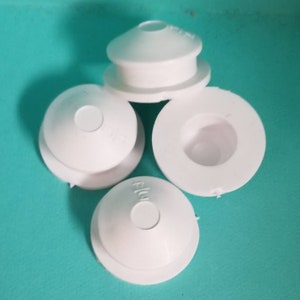 May include: A collection of white plastic parts, possibly for plumbing or industrial use, are arranged on a teal surface. The parts have a circular design with a central hole and a raised, rounded top. Some parts are whole, while others are cut.