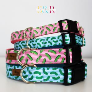 May include: Three dog collars stacked on top of each other. The collars are made of a pink and white striped fabric with a green pickle pattern. The collars have black buckles and silver D-rings.