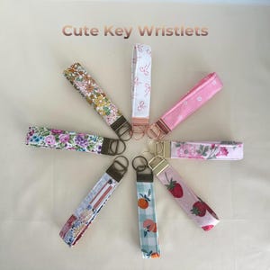 May include: A collection of key wristlets with various floral and fruit patterns. The wristlets feature a variety of colors, including pink, blue, and yellow. Each wristlet has a metal ring and a fabric loop for easy carrying. The text "Cute Key Wristlets" is at the top.