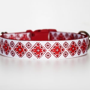 May include: White dog collar with a red geometric pattern. The collar features a red buckle and a silver metal D-ring for leash attachment. The pattern includes red crosses and diamond shapes.
