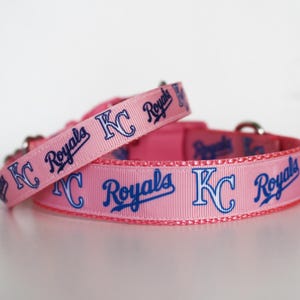 May include: Two pink dog collars with blue and white "KC Royals" text and logo. The collars are made of a soft, pink fabric.