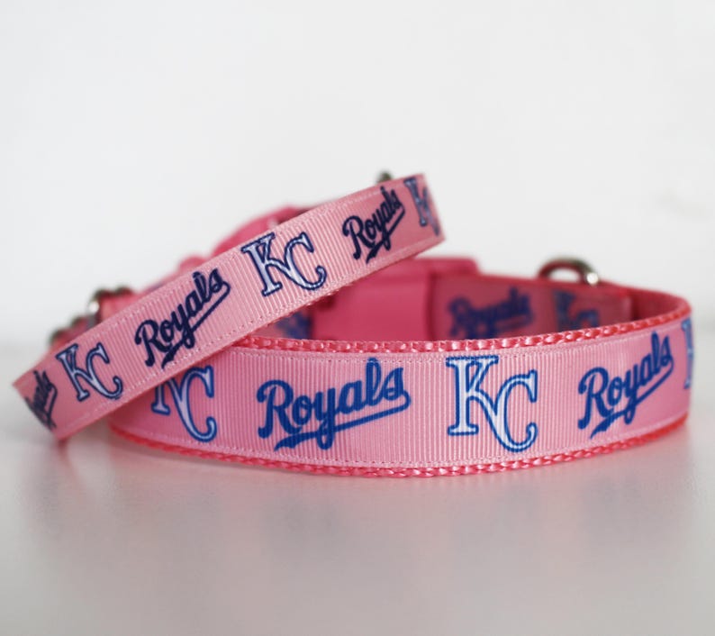 May include: Two pink dog collars with blue and white "KC Royals" text and logo. The collars are made of a soft, pink fabric.