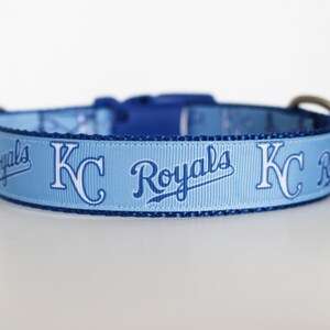 May include: A light blue dog collar with a white and blue Kansas City Royals logo. The collar has a blue and white pattern with the words "Royals" and "KC" repeated.