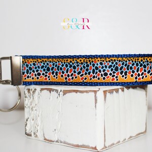 May include: A blue key fob with a patterned design featuring orange, black, and blue spots on a gold background. The key fob has a silver metal clasp and key ring. The text "S O D R" is in the upper left corner.