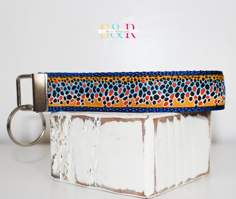 May include: A blue key fob with a patterned design featuring orange, black, and blue spots on a gold background. The key fob has a silver metal clasp and key ring. The text "S O D R" is in the upper left corner.