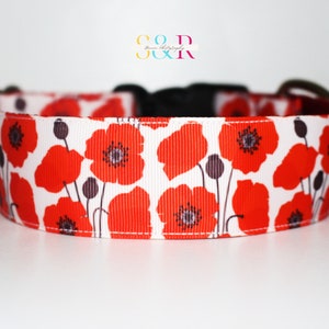 May include: A white dog collar with a red poppy floral print. The collar has a black buckle and a silver D-ring.