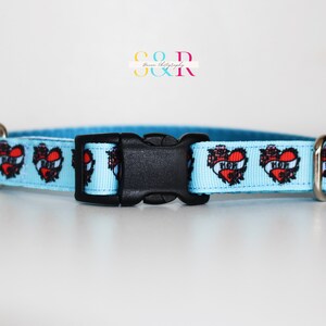 May include: A light blue dog collar with a repeating pattern of red hearts with black outlines and the word "MOM" inside. The collar has a black plastic buckle and silver metal D-rings.