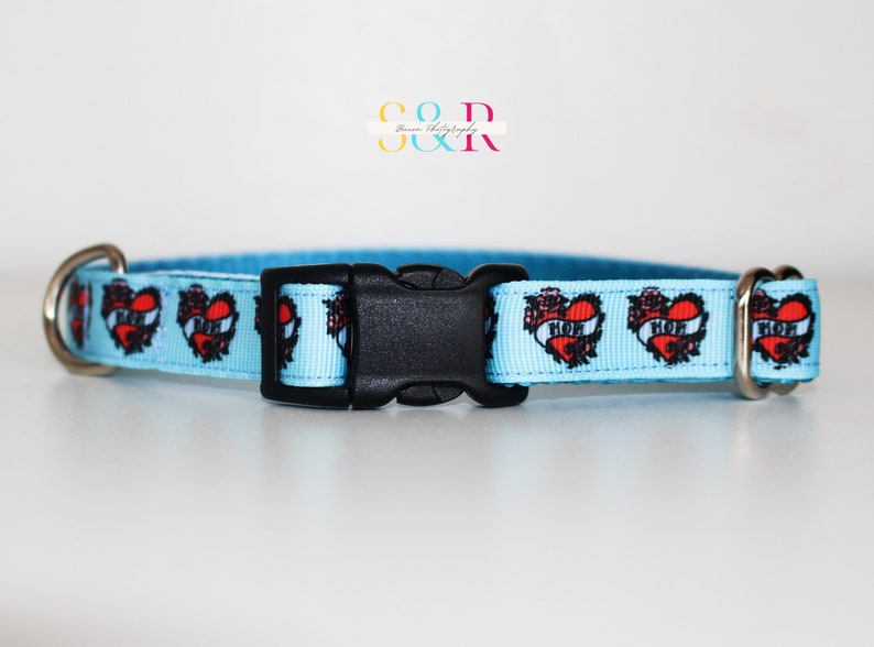May include: A light blue dog collar with a repeating pattern of red hearts with black outlines and the word "MOM" inside. The collar has a black plastic buckle and silver metal D-rings.