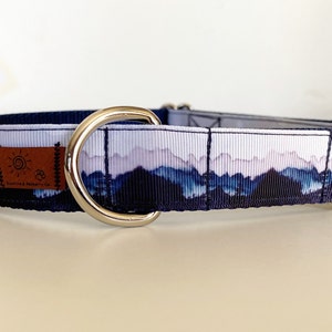 K&ouml;nnte beinhalten: Ein blaues und wei&szlig;es Hundehalsband mit einer Bergszene und einem silbernen D-Ring. Das Halsband hat einen braunen Lederfleck mit dem Text "Footprints & Ruffery Co."