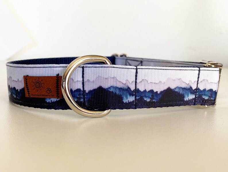 K&ouml;nnte beinhalten: Ein blaues und wei&szlig;es Hundehalsband mit einer Bergszene und einem silbernen D-Ring. Das Halsband hat einen braunen Lederfleck mit dem Text "Footprints & Ruffery Co."