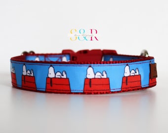 May include: A dog collar featuring a repeating pattern of Snoopy on his doghouse against a light blue background. The collar has a red trim and a red plastic buckle. A small brown leather tag is attached to the collar.