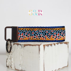 May include: A blue key fob with a brown, orange, black, and blue pattern. The key fob has a bronze-colored metal clasp and key ring. The key fob is sitting on a white, distressed wooden block.