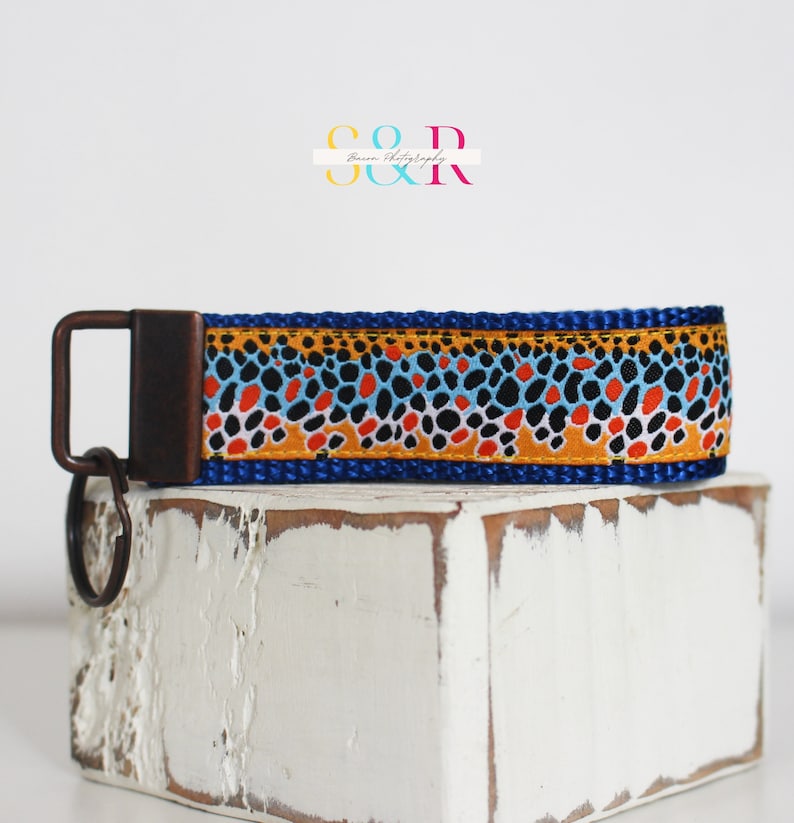 May include: A blue key fob with a brown, orange, black, and blue pattern. The key fob has a bronze-colored metal clasp and key ring. The key fob is sitting on a white, distressed wooden block.