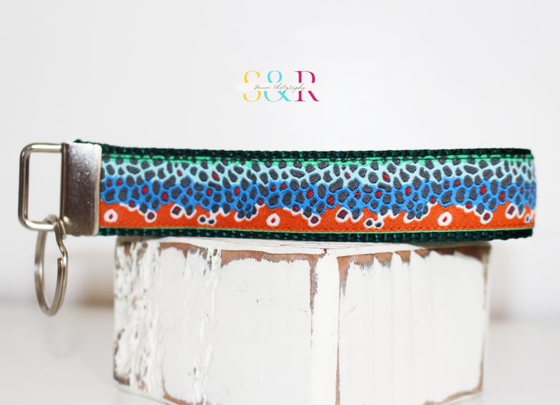 May include: Key fob wrist strap with a fish scale pattern in shades of blue, orange, and gray. The strap is attached to a silver-colored metal key ring and clasp. The design is reminiscent of a trout.