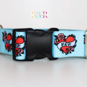 May include: A light blue dog collar with a white background and a repeated pattern of red hearts with the word "MOM" in a banner. The collar has a black plastic buckle and silver metal D-ring.