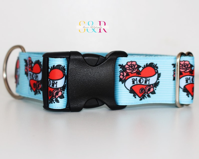 May include: A light blue dog collar with a white background and a repeated pattern of red hearts with the word "MOM" in a banner. The collar has a black plastic buckle and silver metal D-ring.