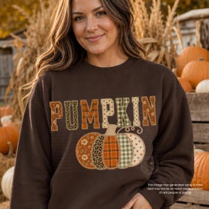 May include: A dark brown crewneck sweatshirt featuring the word PUMPKIN in large, blocky letters with various patterns including floral, plaid, and leopard print. Below the text is a large pumpkin graphic composed of matching patterned segments.