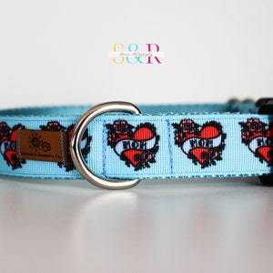 May include: Light blue dog collar with a repeating pattern of red hearts with the word "MOM" inside. The hearts are surrounded by black outlines and floral designs. The collar has a silver D-ring and a black plastic buckle.