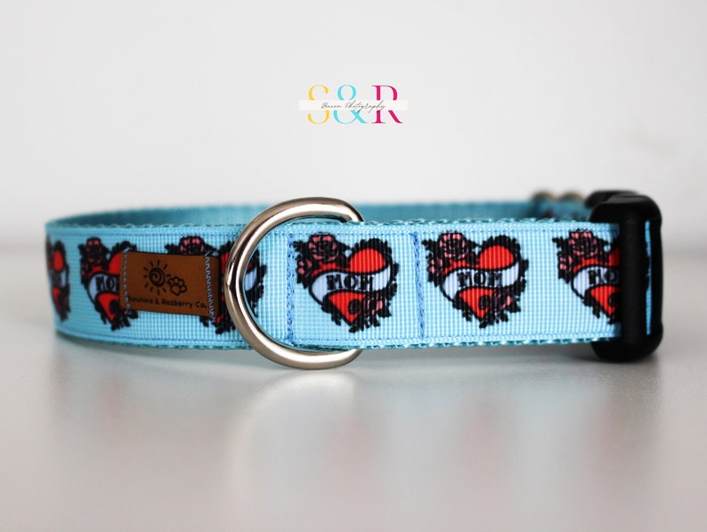 May include: Light blue dog collar with a repeating pattern of red hearts with the word "MOM" inside. The hearts are surrounded by black outlines and floral designs. The collar has a silver D-ring and a black plastic buckle.