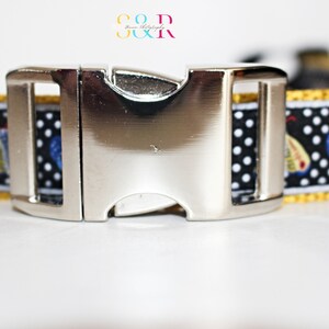 May include: A black and white dog collar with a silver buckle. The collar has a white polka dot pattern and a yellow trim. The collar features a repeating pattern of blue and yellow butterflies.